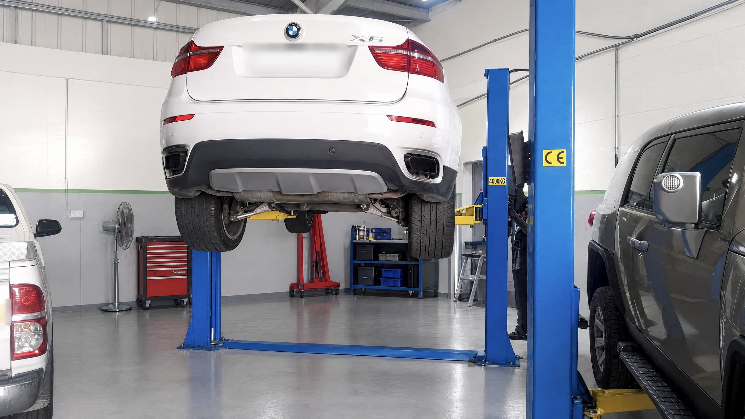 White BMW X6 raised on a blue two-post lift, seen from behind with its wheels hanging and the exhaust and rear suspension exposed, between two other cars in the workshop bays.